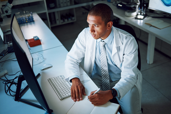male in lab coat sitting at a computer writing in a notebook
